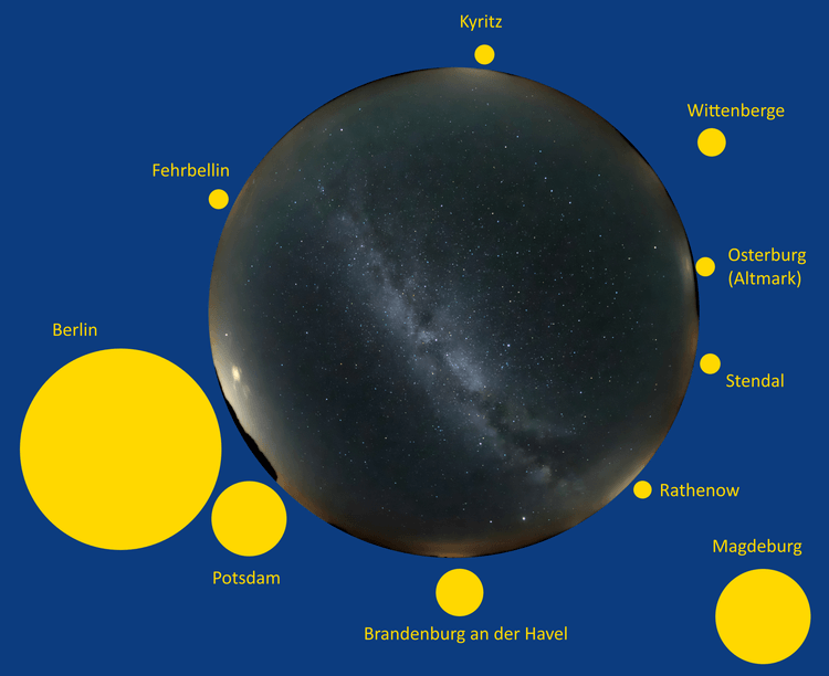 Hemisphärischer abendlicher Frühlingsnachthimmel mit der Milchstraße (in der rechten Bildhälfte) im Internationalen Sternenpark Westhavelland bei zwar wolkenlosem, aber leicht dunstigem Himmel während der astronomischen Dämmerung ohne Mondlicht. Rings herum ist am Horizont die in der Troposphäre durch Streulicht hervorgerufene Lichtverschmutzung der umliegenden Ortschaften zu sehen, im Osten (links) die Lichtglocke vom 50 bis 90 Kilometer entfernten Berlin.